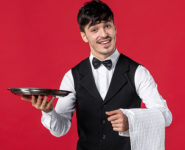 Front view of young male waiter in a uniform with butterfly on neck and holding tray towel on red background