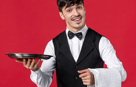 Front view of young male waiter in a uniform with butterfly on neck and holding tray towel on red background