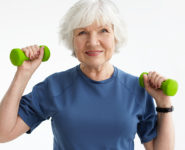 People, age, sports and active lifestyle concept. Picture of happy positive mature retired woman in t-shirt doing exercise with free weights in gym. Excited senior female training with dumbbells